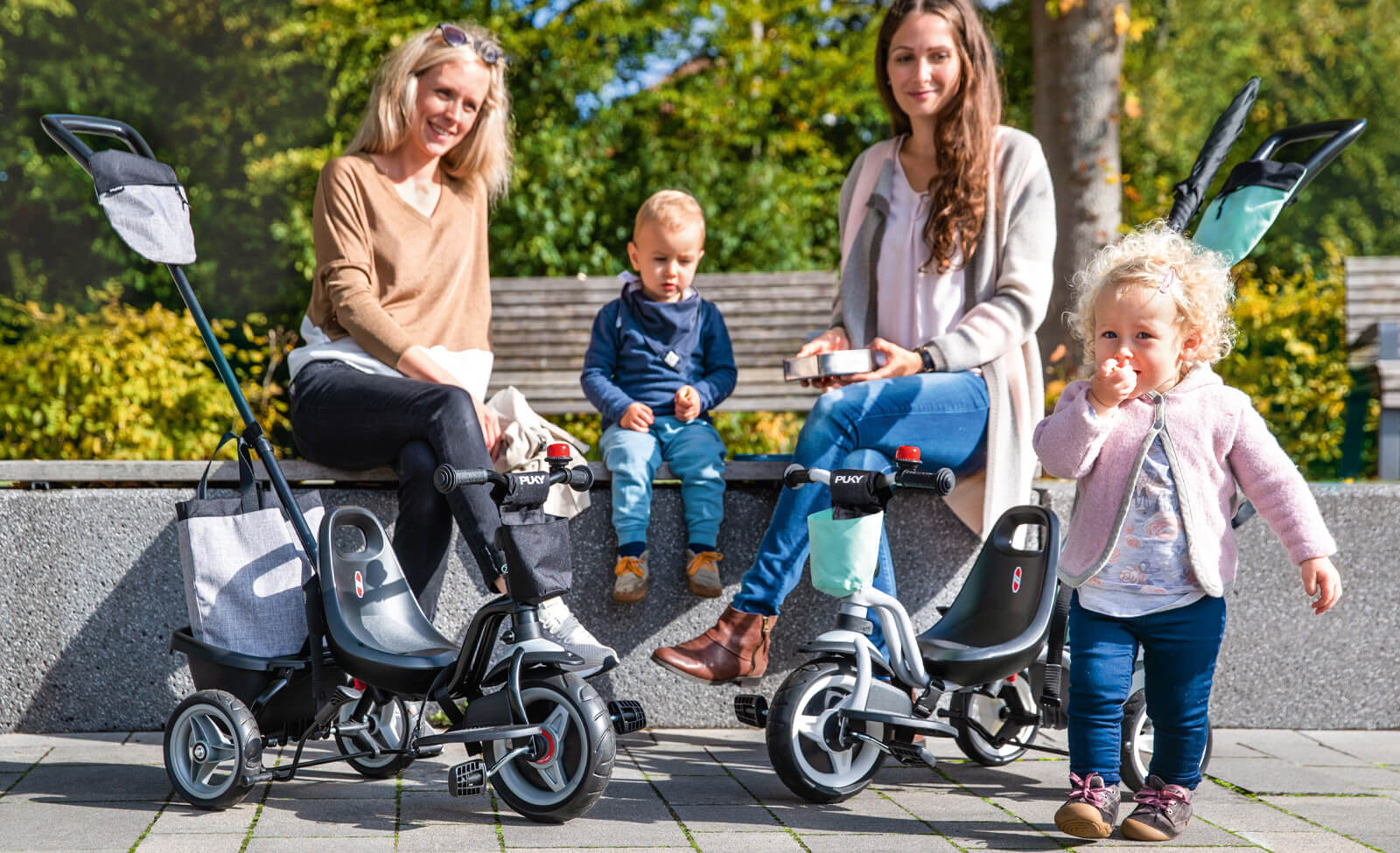 Kinder auf Puky Dreirädern mit Eltern, draußen auf Steinbank im Park sitzend, sonniges Wetter.