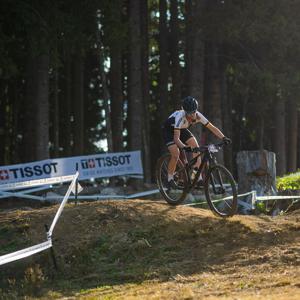 Mountainbiker auf XC-Kurs, fährt über eine Erdanhöhe im Wald, Tissot-Banner im Hintergrund, sonniges Wetter.