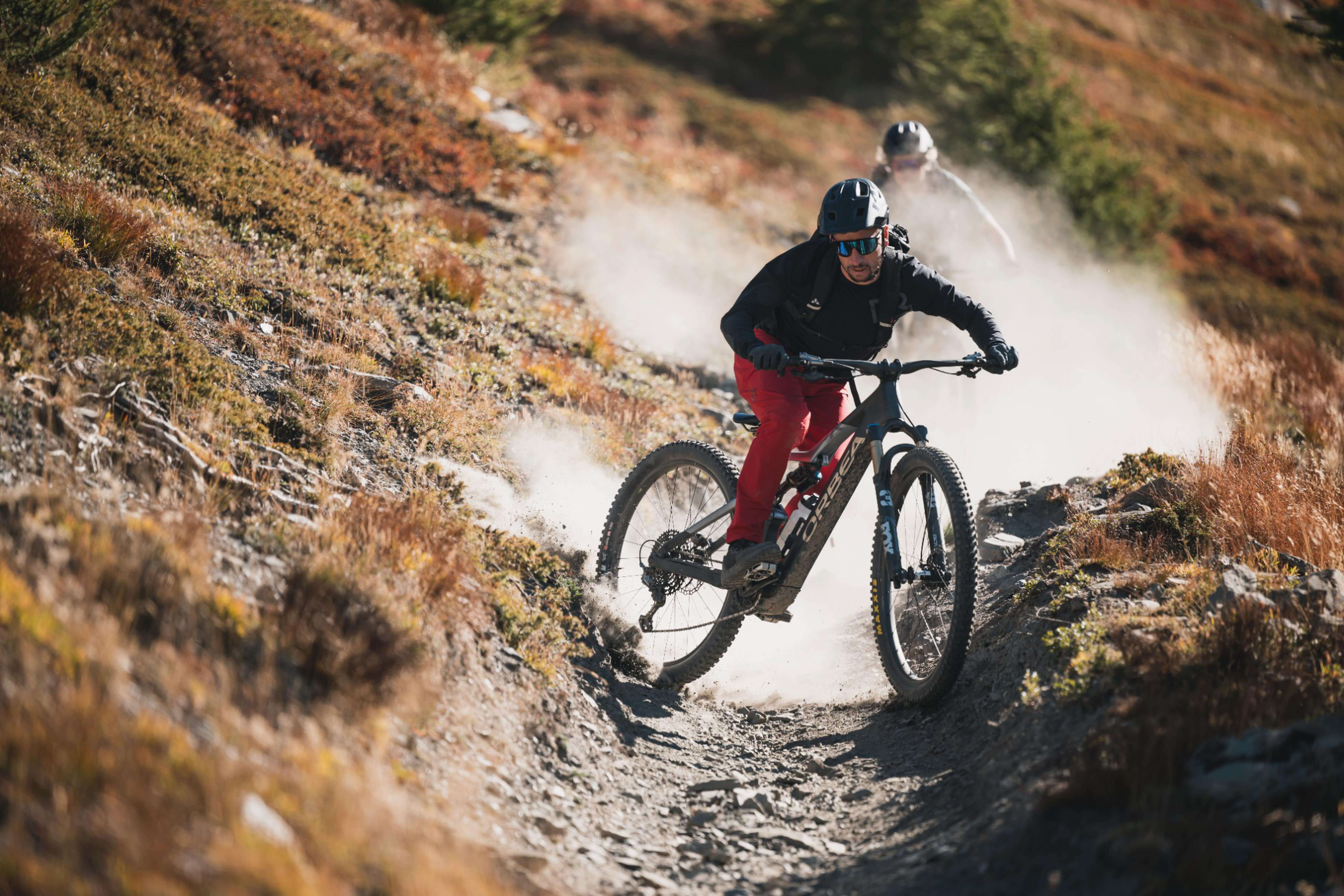 Zwei Mountainbiker fahren auf einem staubigen Trail bergab. Im Vordergrund ein Orbea E-Mountainbike.