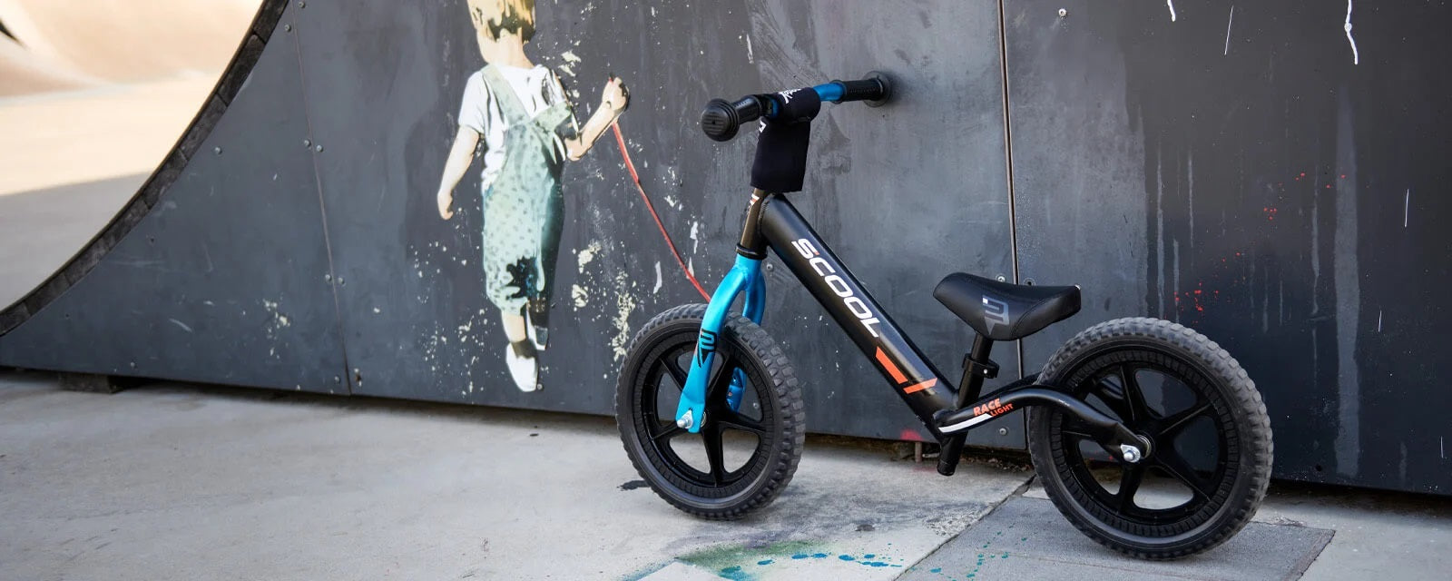 Laufrad für Kinder vor Halfpipe, Modell S'COOL Race mit schwarzem Rahmen und blauen Details. Ideal für den Skatepark.