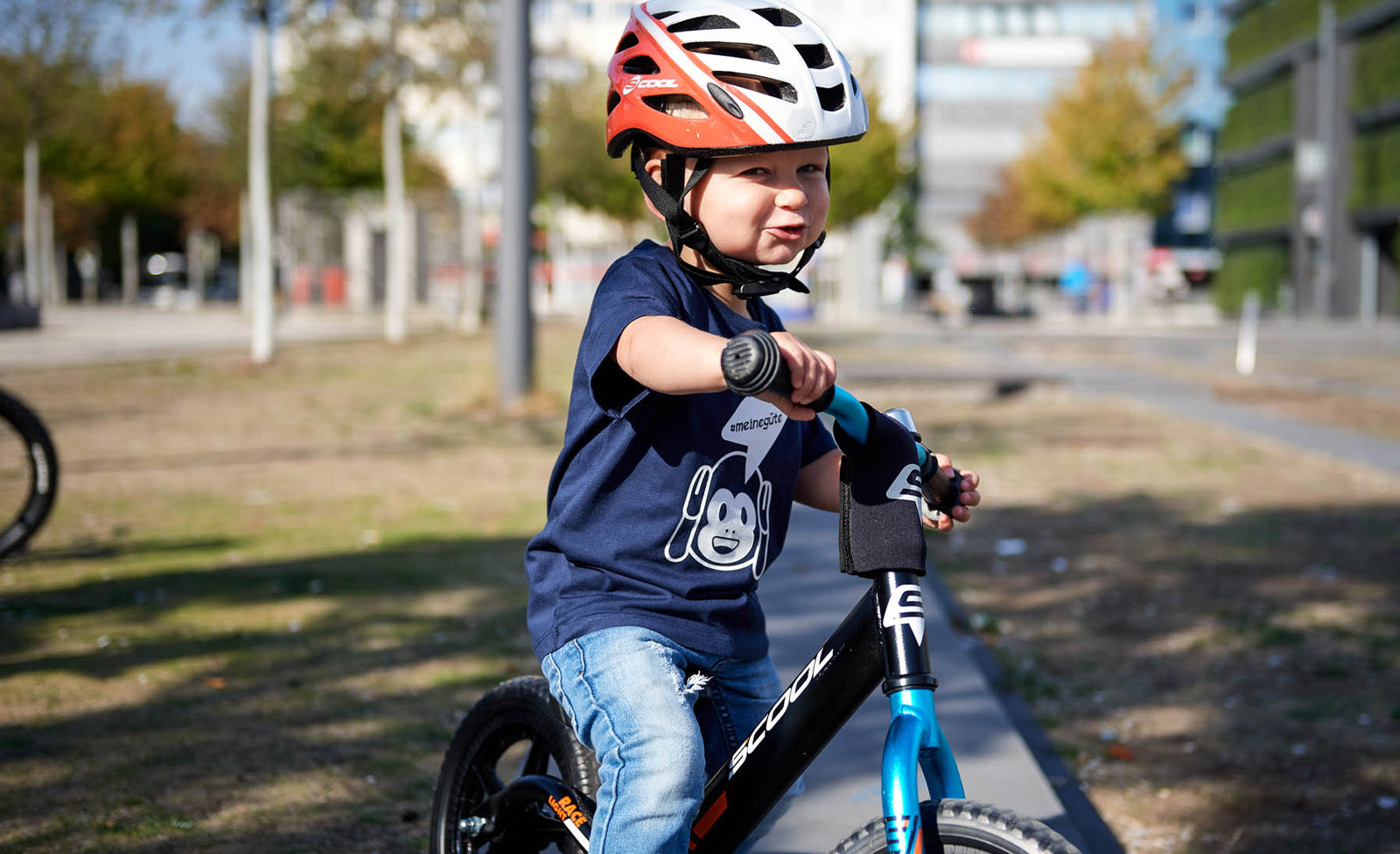 Kind mit S'cool-Laufrad und Helm im Park auf Asphaltweg. Geeignet für junge Radfahrer zur Sicherheit und Balance.
