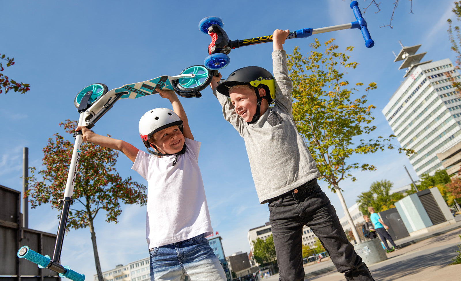 Zwei Kinder mit Helmen heben fröhlich ihre Scooter in die Luft, draußen auf einem Platz, sonniger Tag.