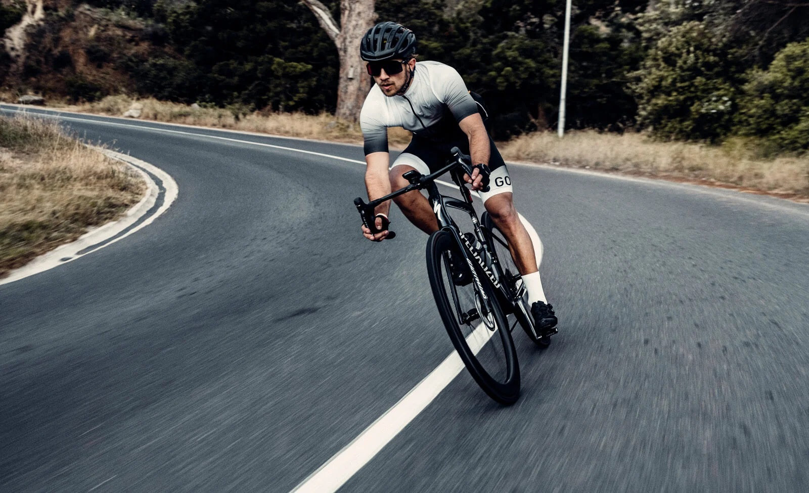 Radfahrer in Kurve mit Specialized-Rennrad und GORE-Bekleidung auf Landstraße. Helm und Sonnenbrille für Sicherheit.