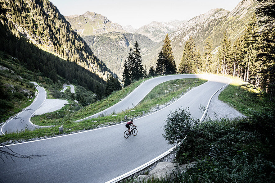 Radfahrer auf gewundener Bergstraße, umgeben von Wald und Bergen. Ideal für Radsport im Freien. Marke nicht erkennbar.