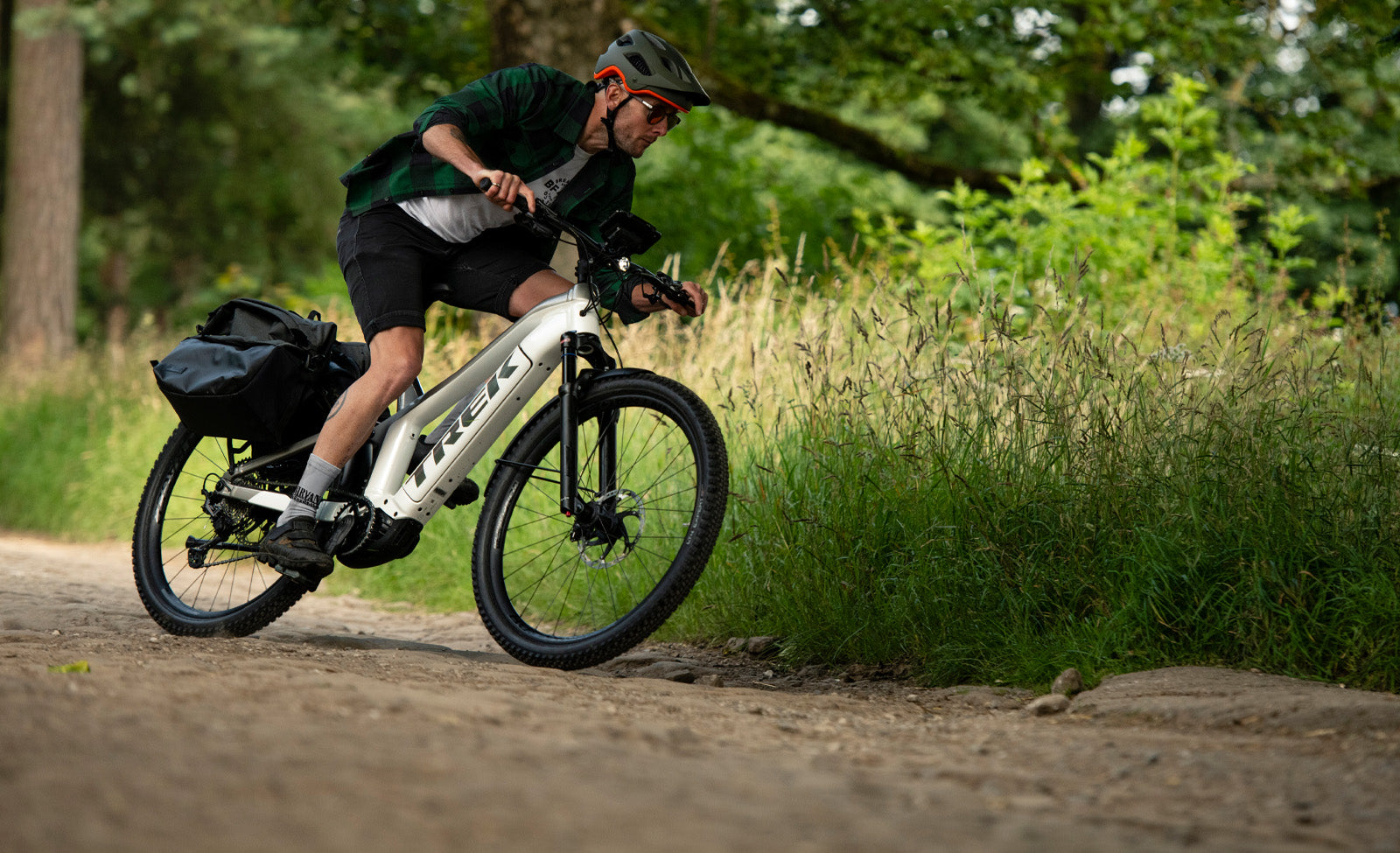 Person auf Trek E-MTB Hardtail fährt in Waldweg, trägt Helm und sportliche Kleidung, hat seitliche Gepäcktaschen.