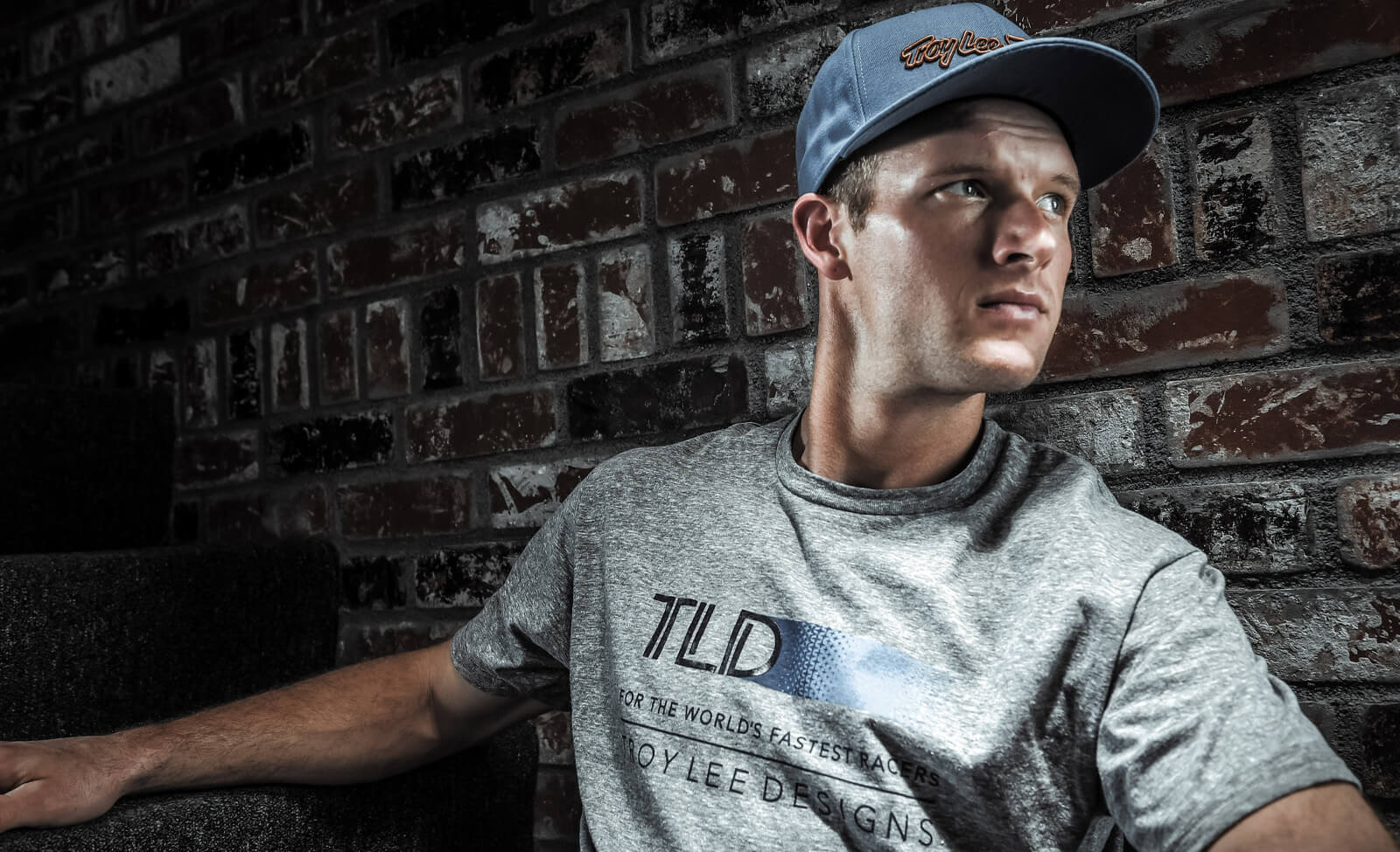 Mann mit Troy Lee Designs T-Shirt und Kappe sitzt vor Backsteinwand. Urbaner, sportlicher Look für junge Menschen.
