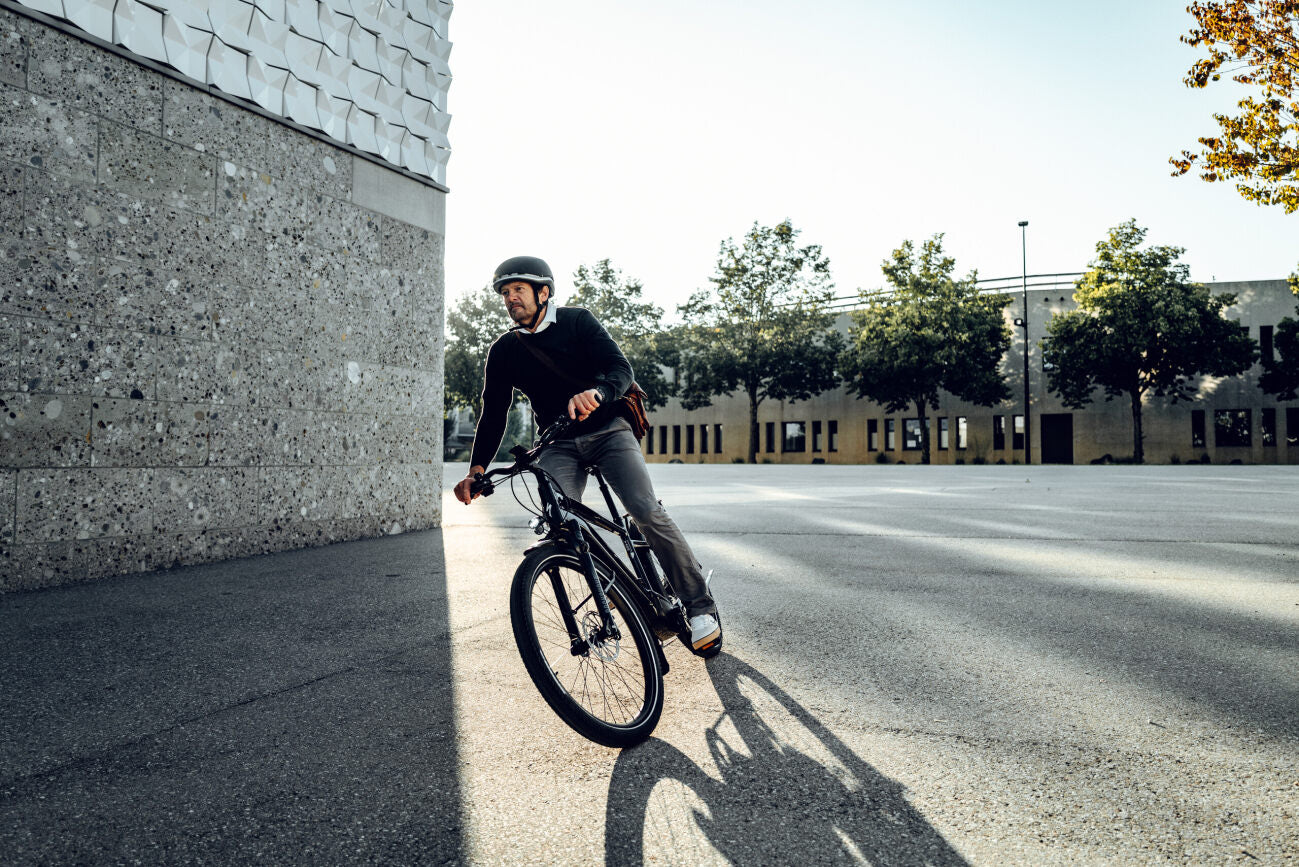 Mann fährt Specialized E-Trekkingrad auf leerem Platz, trägt Helm, moderne Architektur im Hintergrund, sonniger Tag.