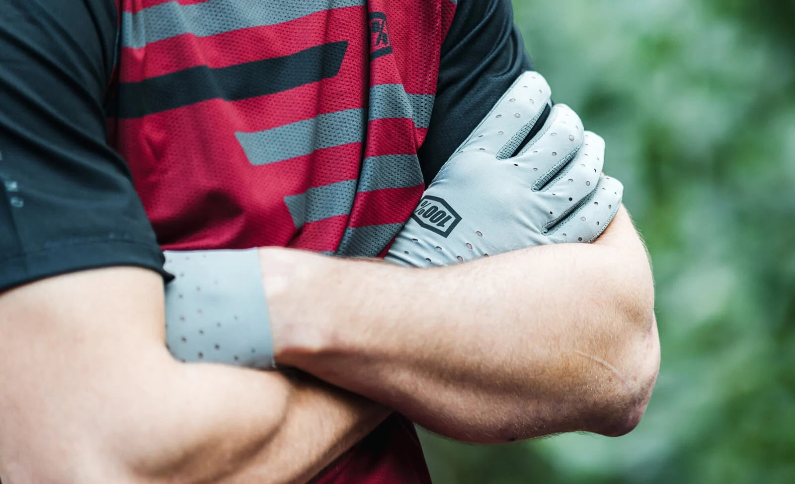 Person trägt graue 100% Fahrrad-Handschuhe und ein rot-schwarzes Trikot, Arme verschränkt vor grünem Hintergrund.