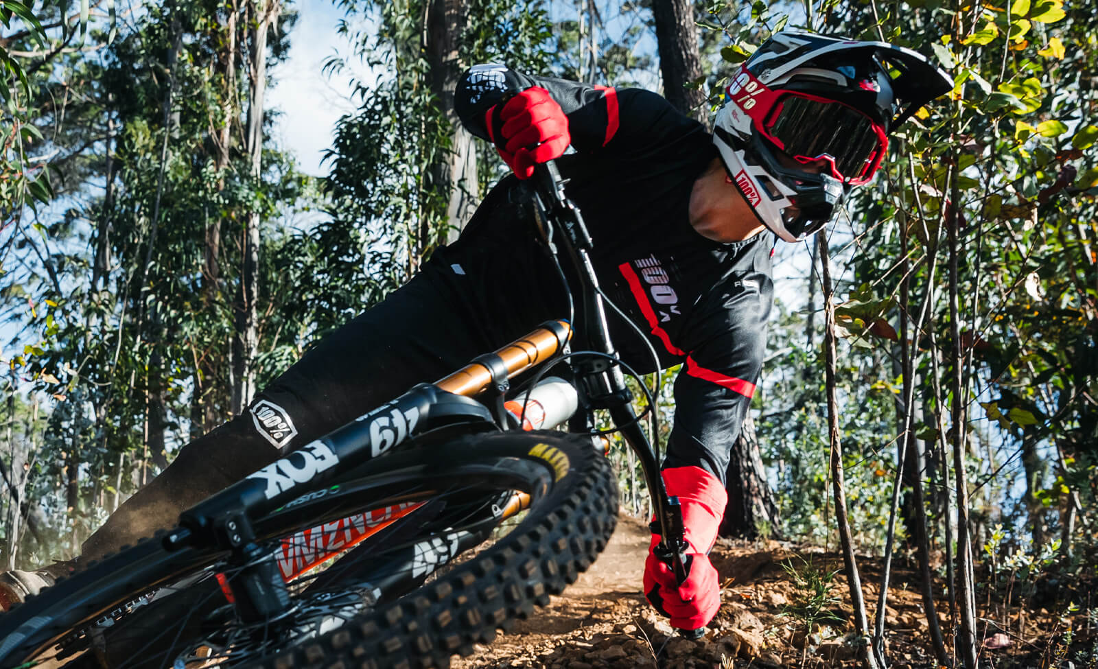 Mountainbiker in 100% Gear fährt steilen Waldweg, trägt Helm und Schutzbrille, geeignet für Downhill-Aktivitäten.