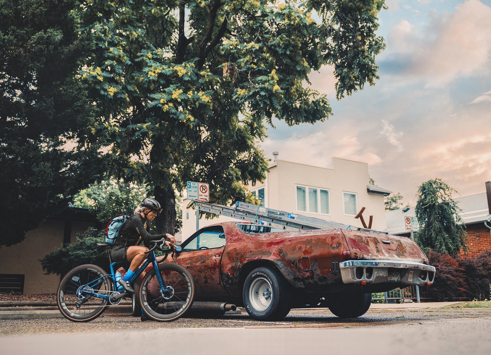 Radfahrerin auf Mondraker Arid Fahrrad neben rostigem Auto, Baum im Hintergrund, urbanes Umfeld.