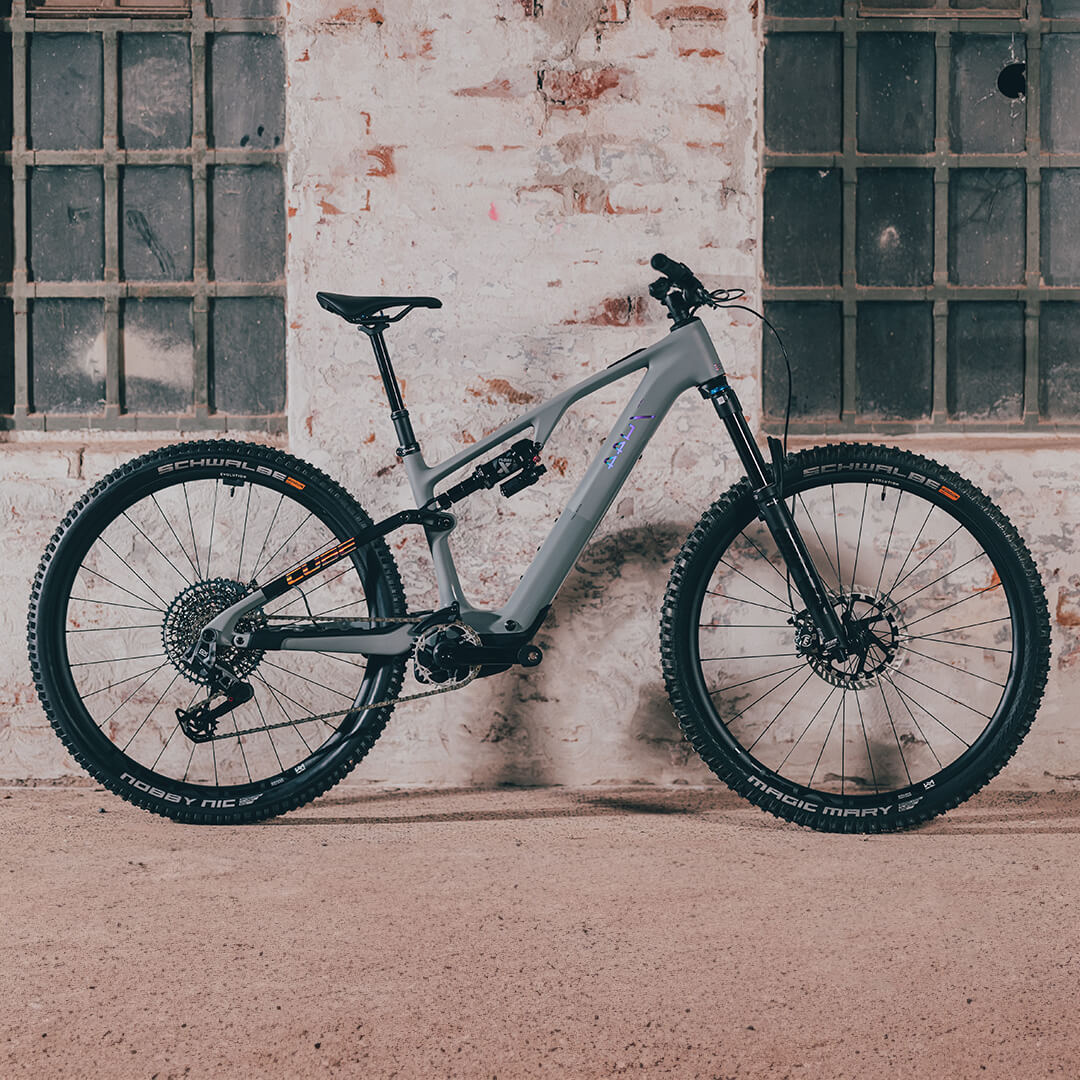 Graues Cube Mountainbike, Modell 2025, mit Vollfederung und Schwalbe-Reifen, steht vor einer alten Backsteinmauer.