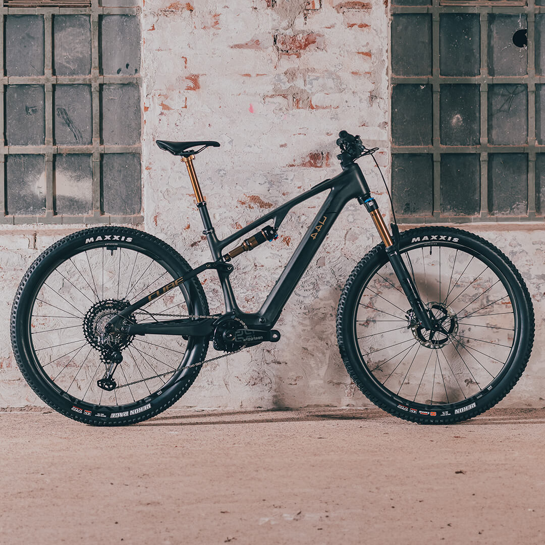 Schwarzes Mountainbike von CUBE, Modell 2025, vor rustikaler Wand, mit goldenen Akzenten und Maxxis-Reifen.