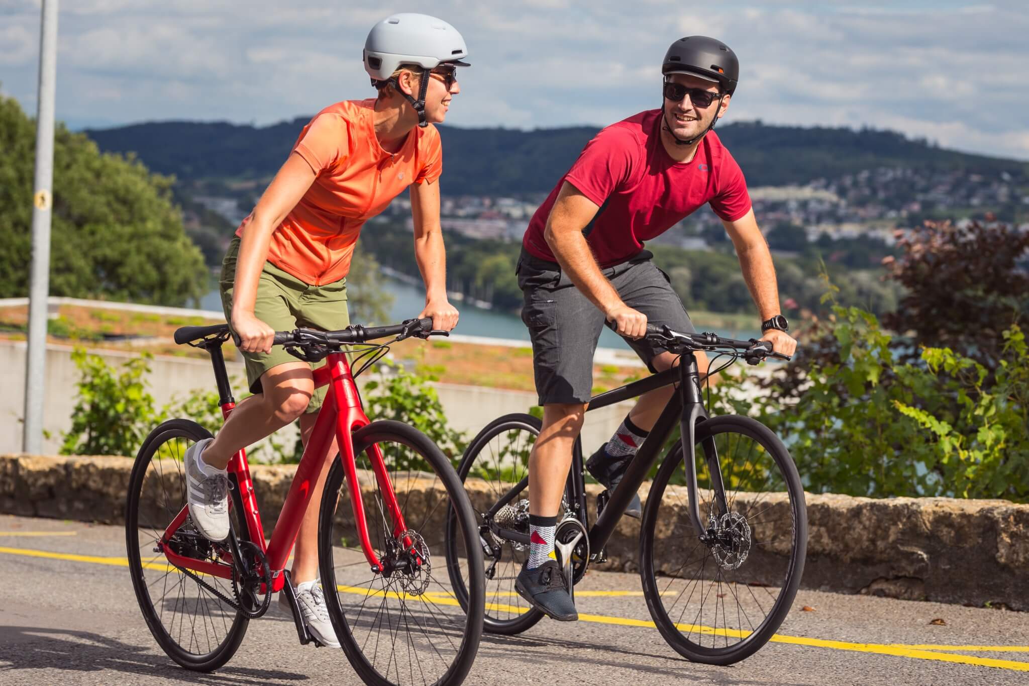 Zwei Personen fahren BMC Alpenchallenge Fahrräder auf einer Straße in ländlicher Umgebung, beide tragen Helme.