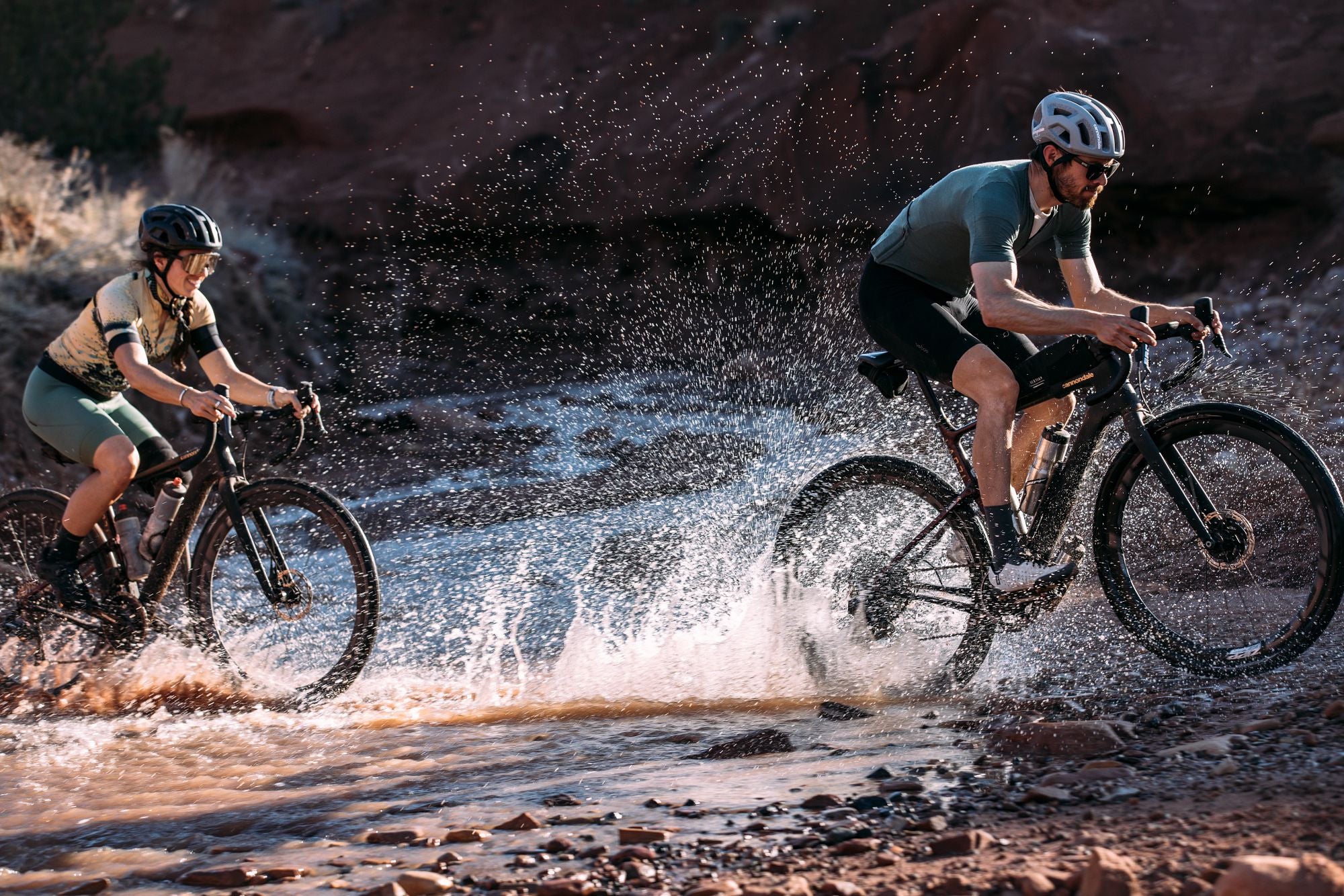 Zwei Radfahrer in Sportkleidung fahren durch flaches Wasser und spritzen Wasser auf einem unbefestigten Weg.