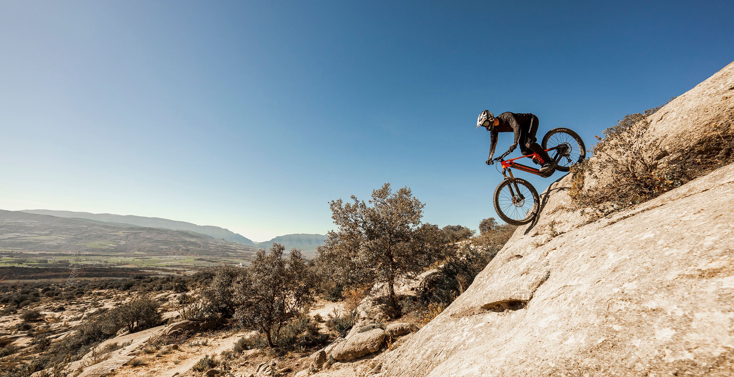 Mountainbiker auf steilem Felsabhang mit Mondraker Crafty E-Mountainbike in wüstenähnlicher Landschaft, blauer Himmel.