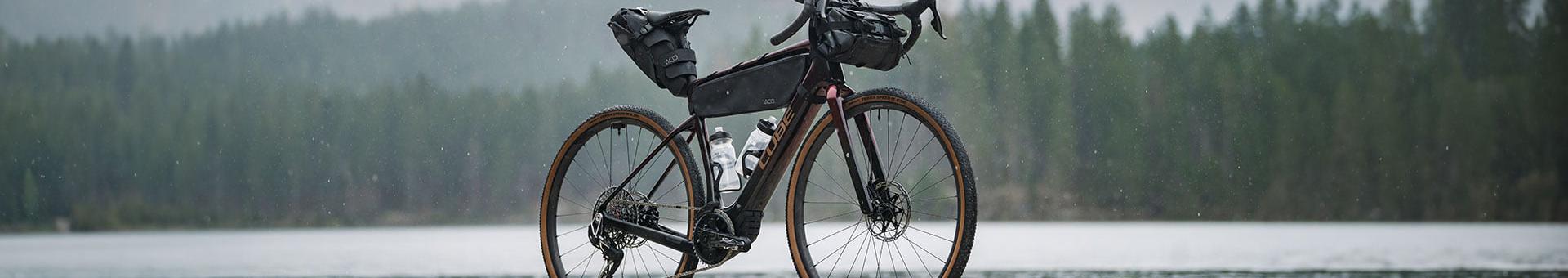 Cube E-Gravelbike mit Taschen für Abenteuerfahrten, seitlich am Seeufer in regnerischer Waldlandschaft fotografiert.