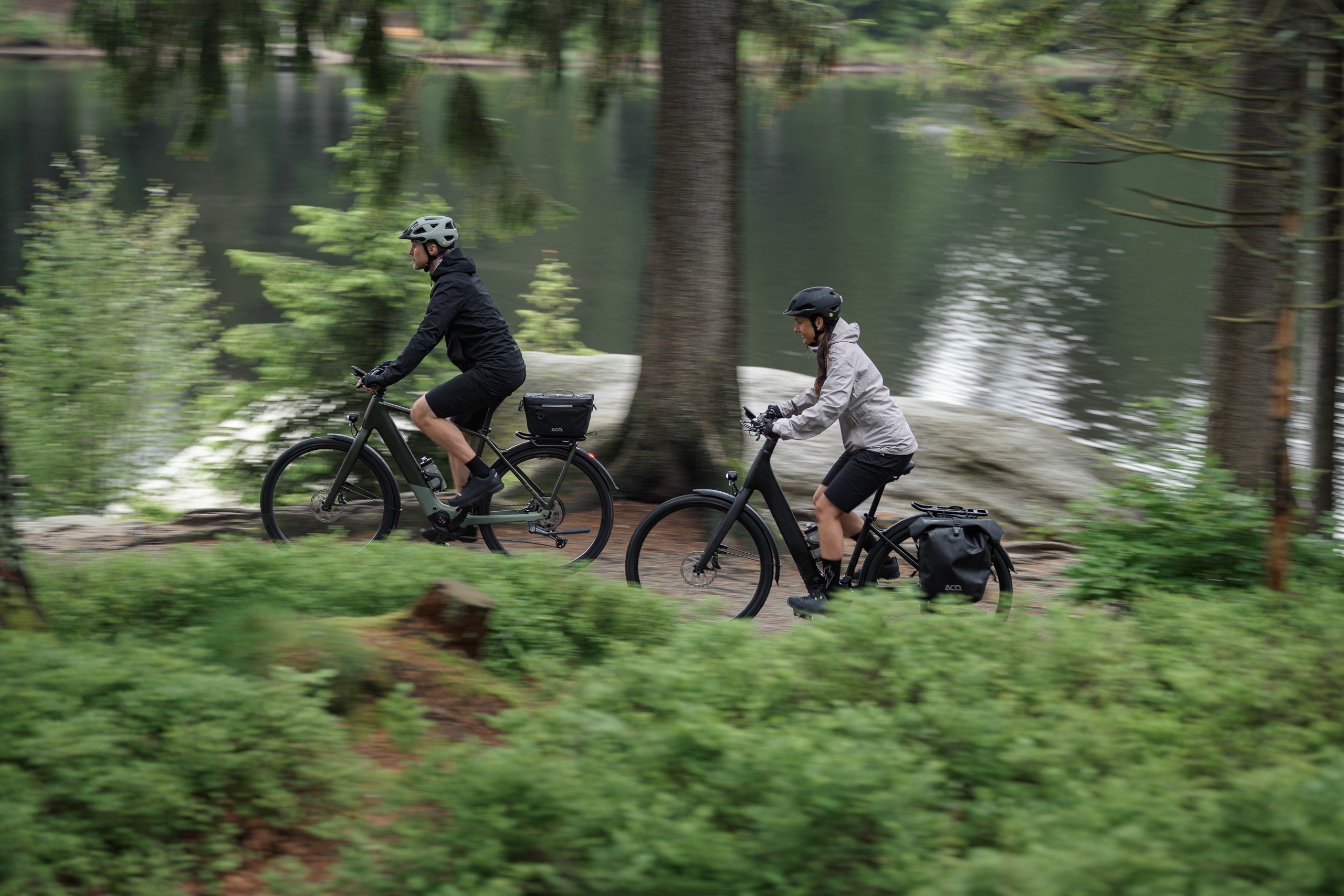Zwei Personen fahren Cube E-Bikes auf einem Waldweg neben einem See, geeignet für Outdoor-Enthusiasten.