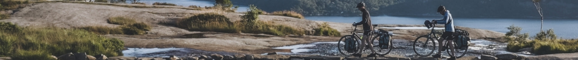 Zwei Personen fahren mit Cube Trekkingrädern entlang eines felsigen Pfades in einer naturnahen Umgebung am Wasser.