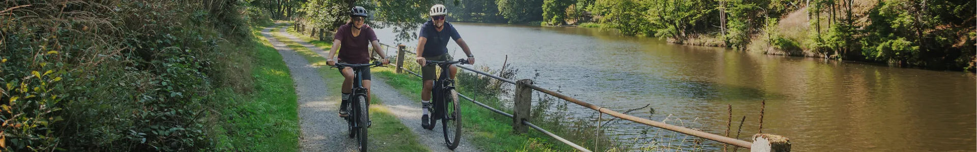 Zwei Personen fahren ein CUBE Reaction Hybrid ONE44 E-Bike auf einem Waldweg entlang eines Flusses.