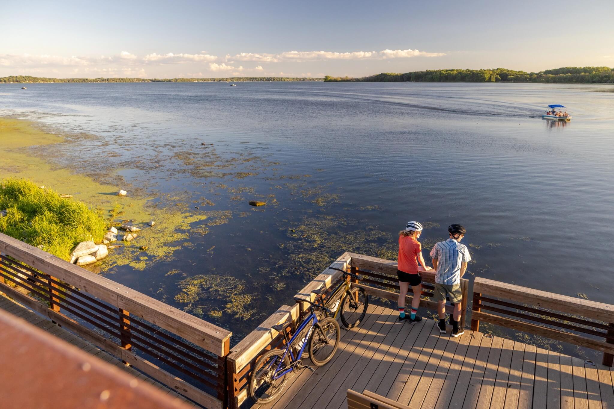 Zwei Personen mit Fahrrädern, die auf einem Steg am ruhigen See pausieren, Sommerbekleidung, Helm, idyllische Natur.