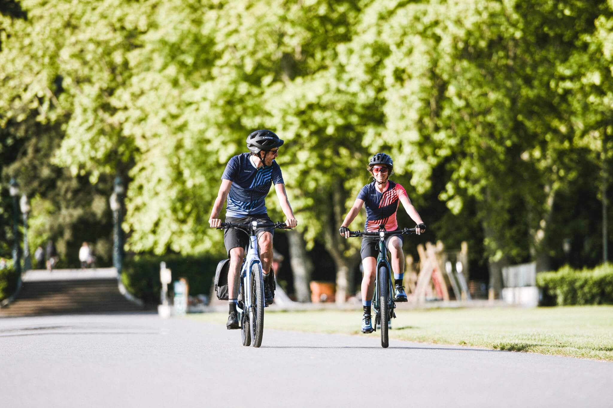 Zwei Radfahrer auf City-Trekkingrädern in einem Park bei Sonnenschein, beide tragen Helme und sportliche Kleidung.