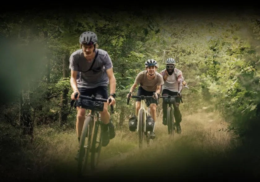Drei Personen fahren mit Gravel-Bikes auf einem Waldweg, alle tragen Helme und haben Fahrradtaschen; Outdoor-Abenteuer.