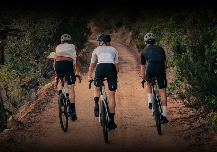 Drei Radfahrer auf einer Schotterstraße im Wald, tragen Shorts und Helme, fahren Gravelbikes.