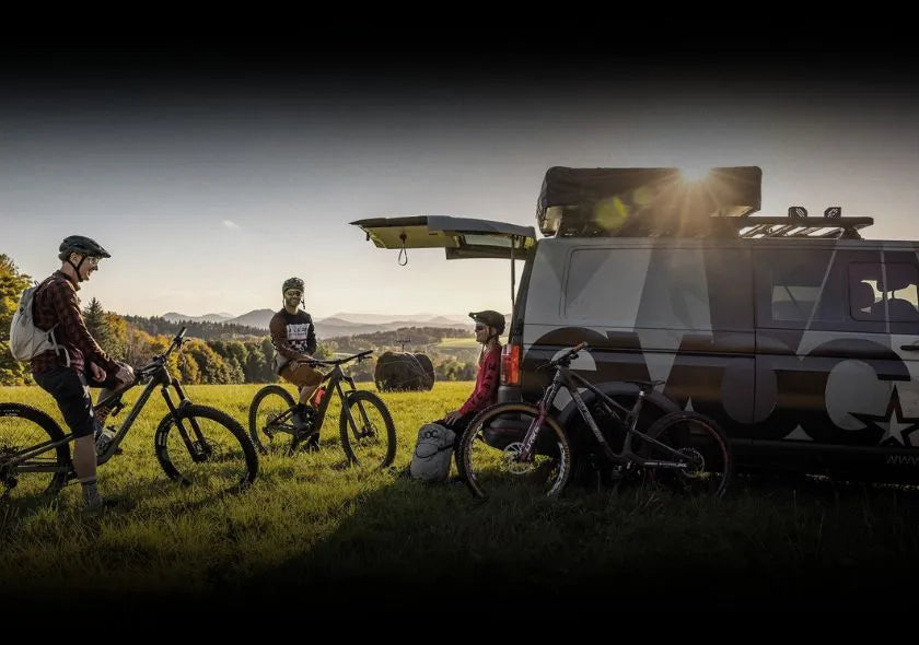 Mountainbiker und Camper-Van auf Wiese in der Abendsonne, zwei Männer auf Fahrrädern, eine Person sitzt am Van.