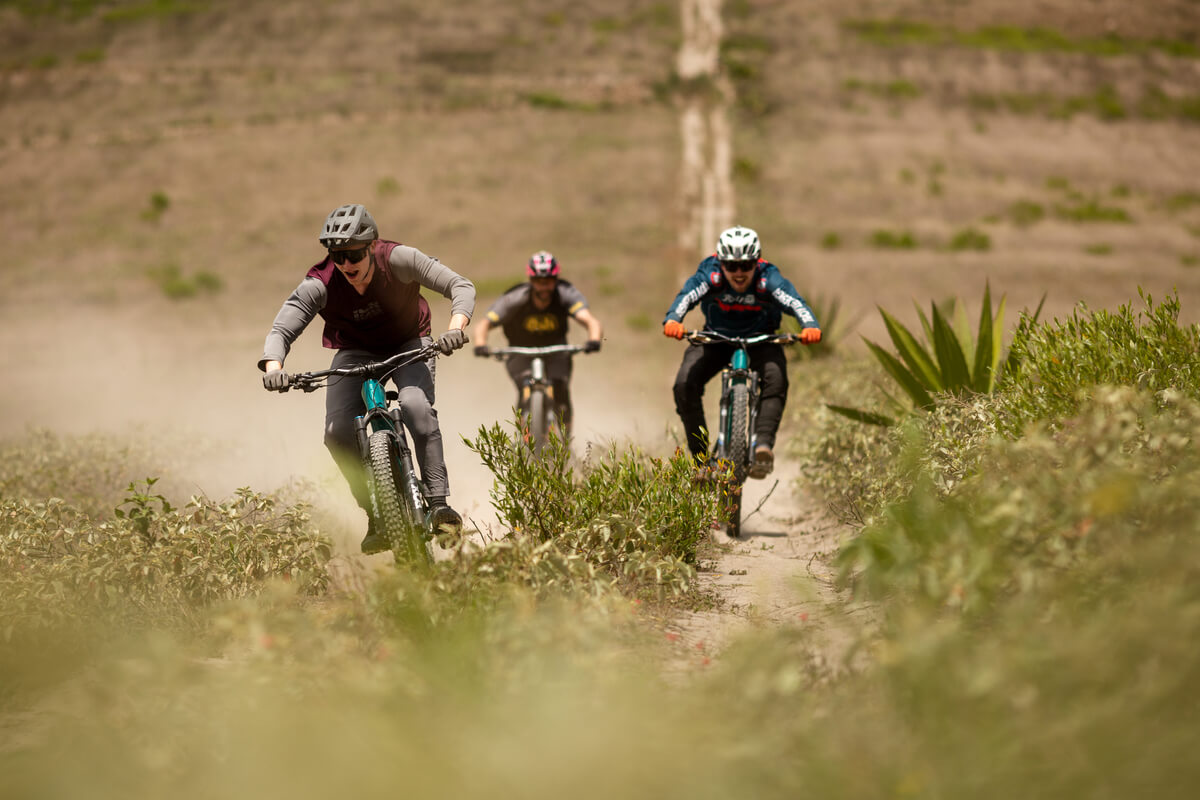Drei Mountainbiker fahren auf einem staubigen Pfad durch hügeliges Gelände, zwei tragen Helme und spezielle Radkleidung.