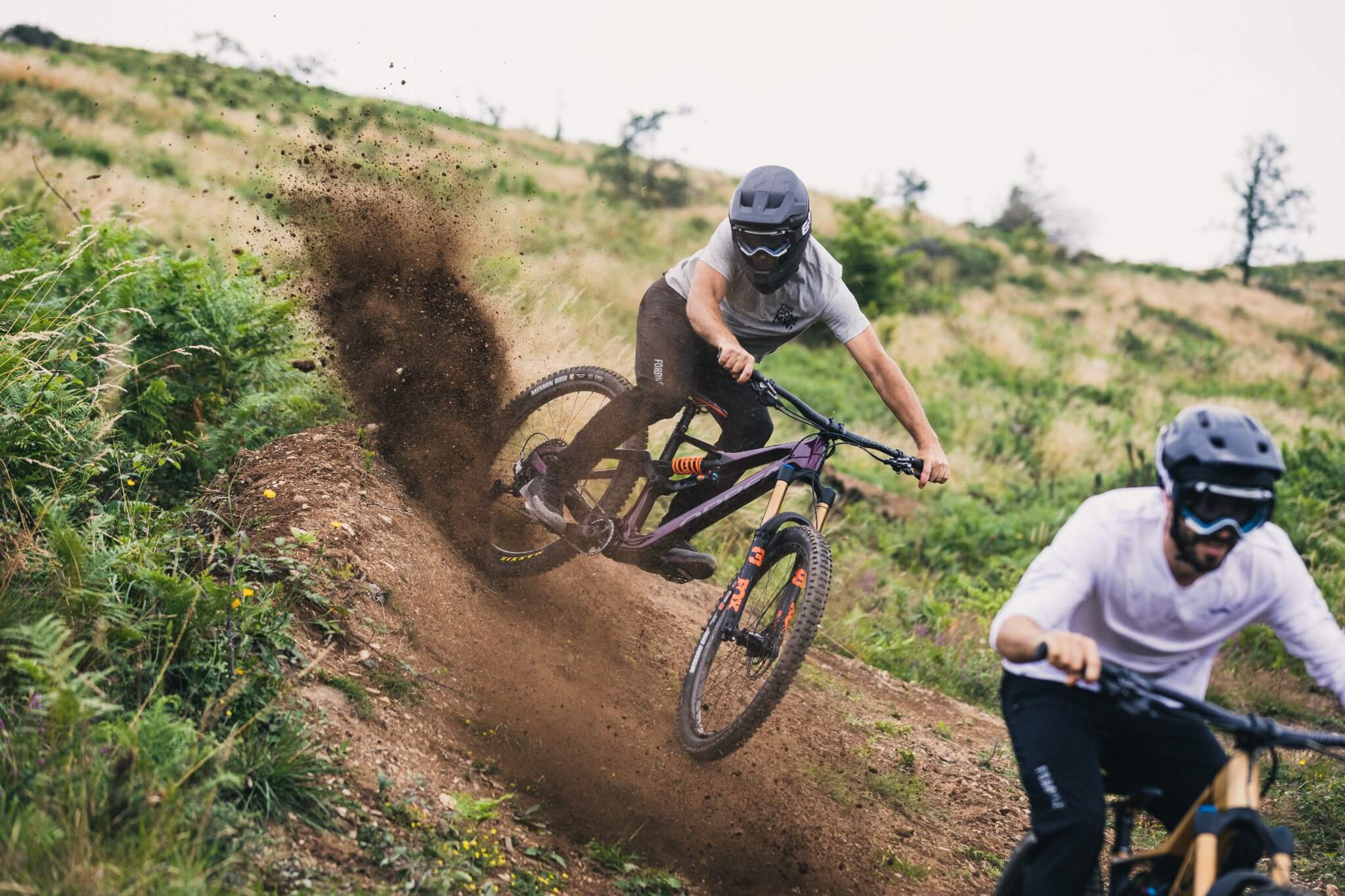 Zwei Mountainbiker fahren mit Helm und Schutzkleidung über einen staubigen Hang in einer grünen Landschaft.