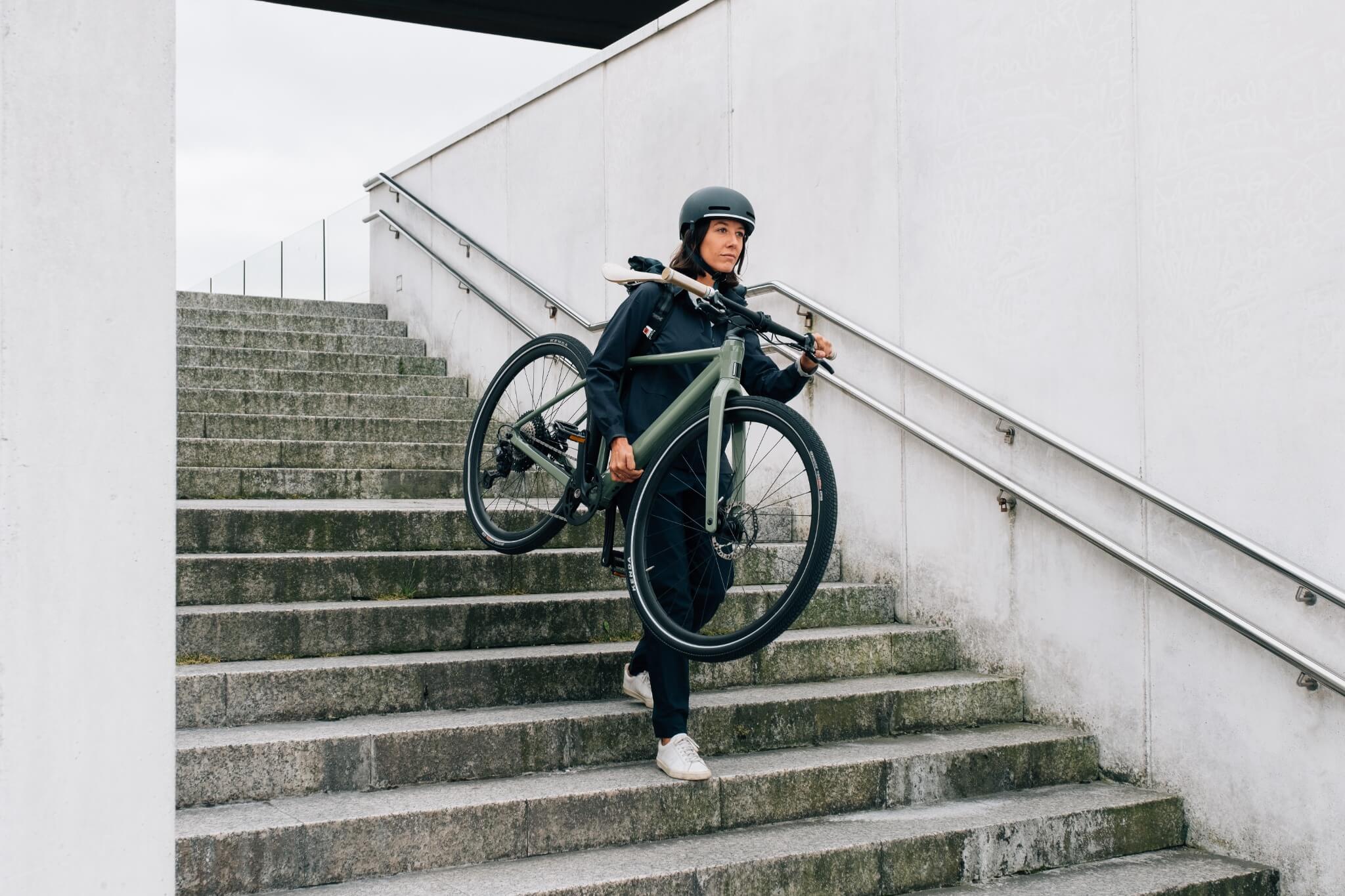 Person trägt ein grünes Stadtrad die Treppe hinunter. Einsatzbereich: Mobilität in der Stadt. Modell: Orbea e-City.