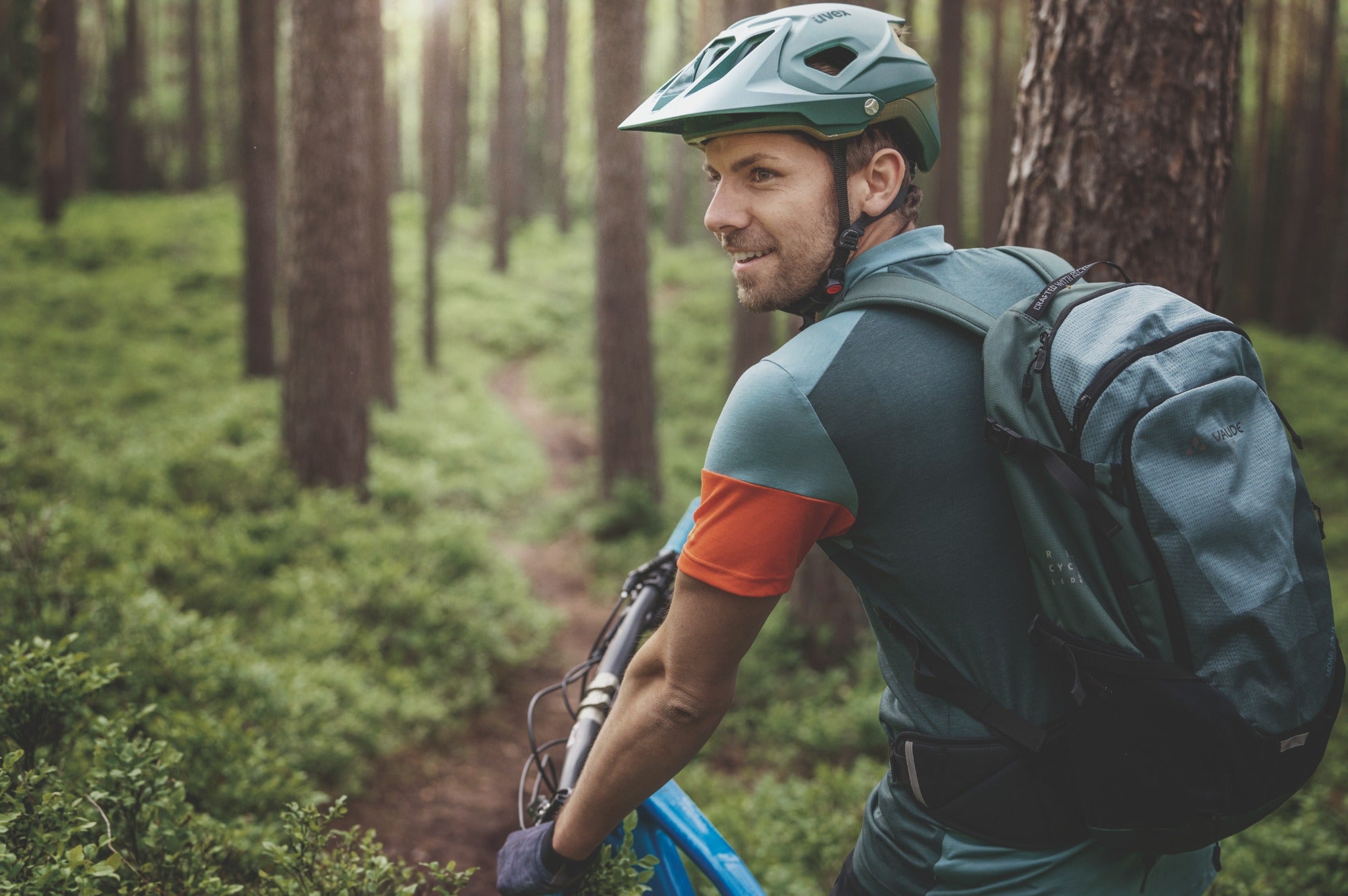 Mann mit Mountainbike, trägt Uvex-Helm und Vaude-Rucksack im Wald, auf einem Trail unterwegs.