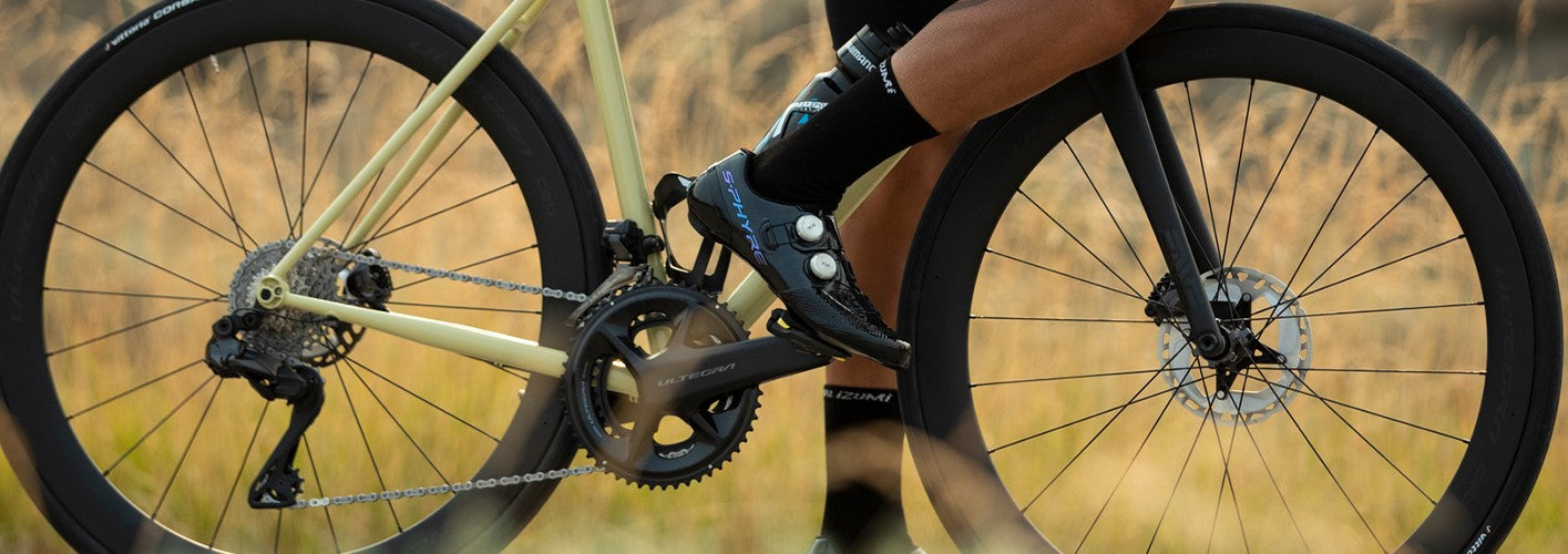 Rennrad mit gelbem Rahmen, Shimano Ultegra Schaltung, Drahtreifen und Fahrer in Radbekleidung auf ländlicher Straße.