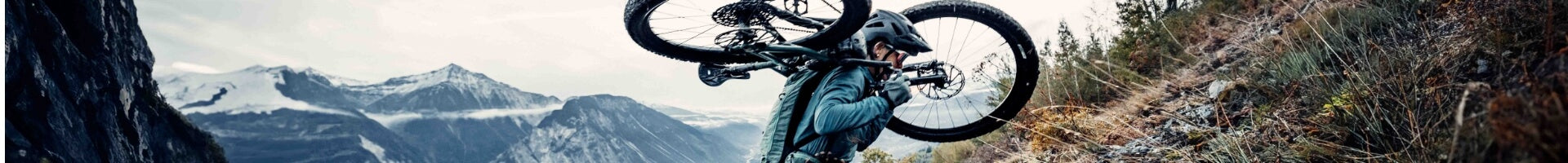 Radfahrer trägt E-Mountainbike bergauf, in alpiner Landschaft, Fokus auf Specialized E-MTB Hardtail, für Offroad-Abenteuer.