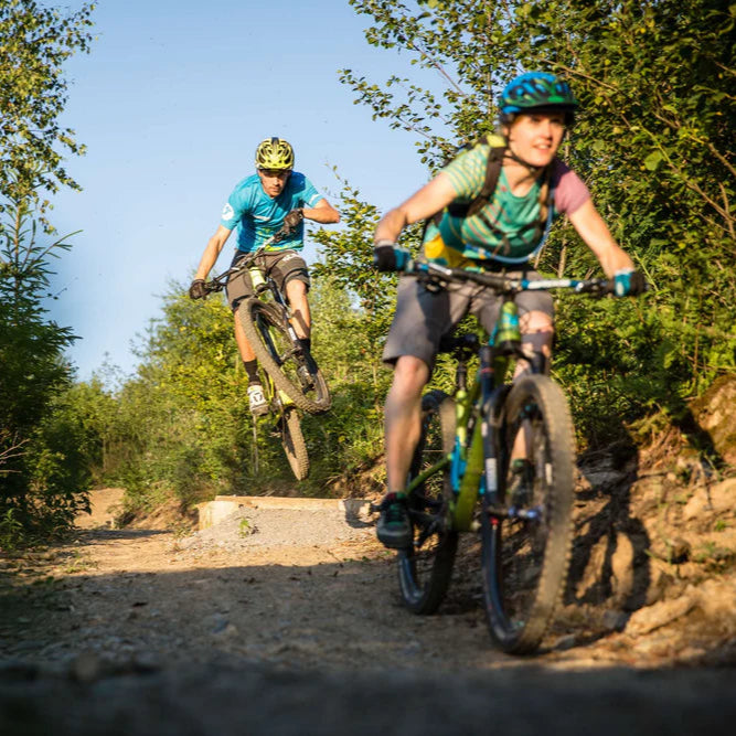 Zwei Personen fahren Mountainbike im Wald, eine springt über eine Rampe.