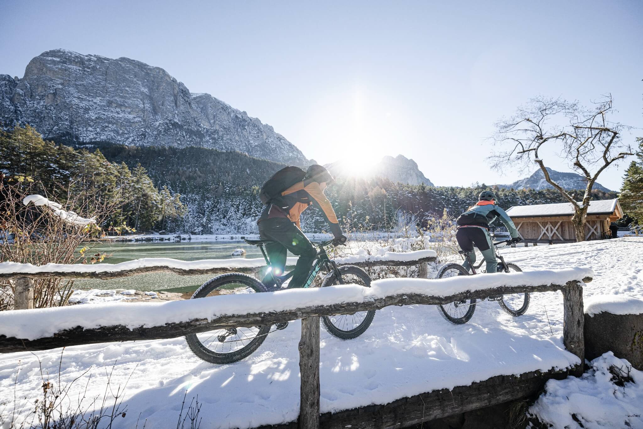 Zwei Personen fahren Mountainbike auf verschneitem Weg vor Bergkulisse und See, eingetaucht in Sonnenlicht.