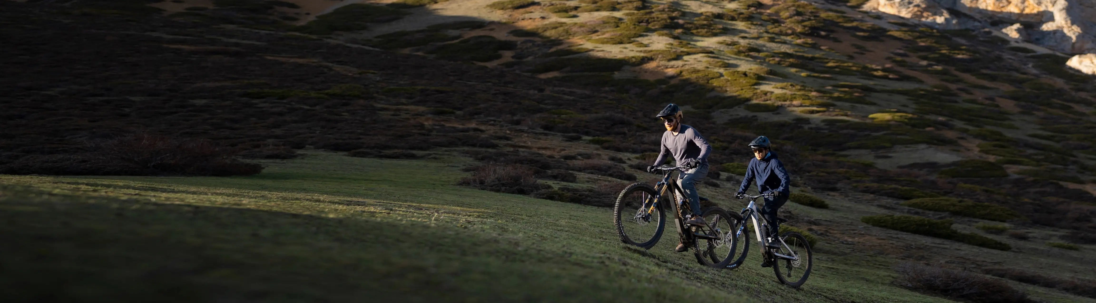 Zwei Personen fahren mit Mountainbikes einen grünen Hügel hinauf; sie tragen Helme und sportliche Kleidung.