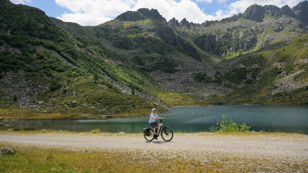 Person fährt ein E-Bike CUBE KATHMANDU HYBRID DELUXE PRO 800
auf einem Schotterweg vor einem Bergsee, umgeben von grüner Landschaft. Ideal für Outdoor-Aktivitäten.