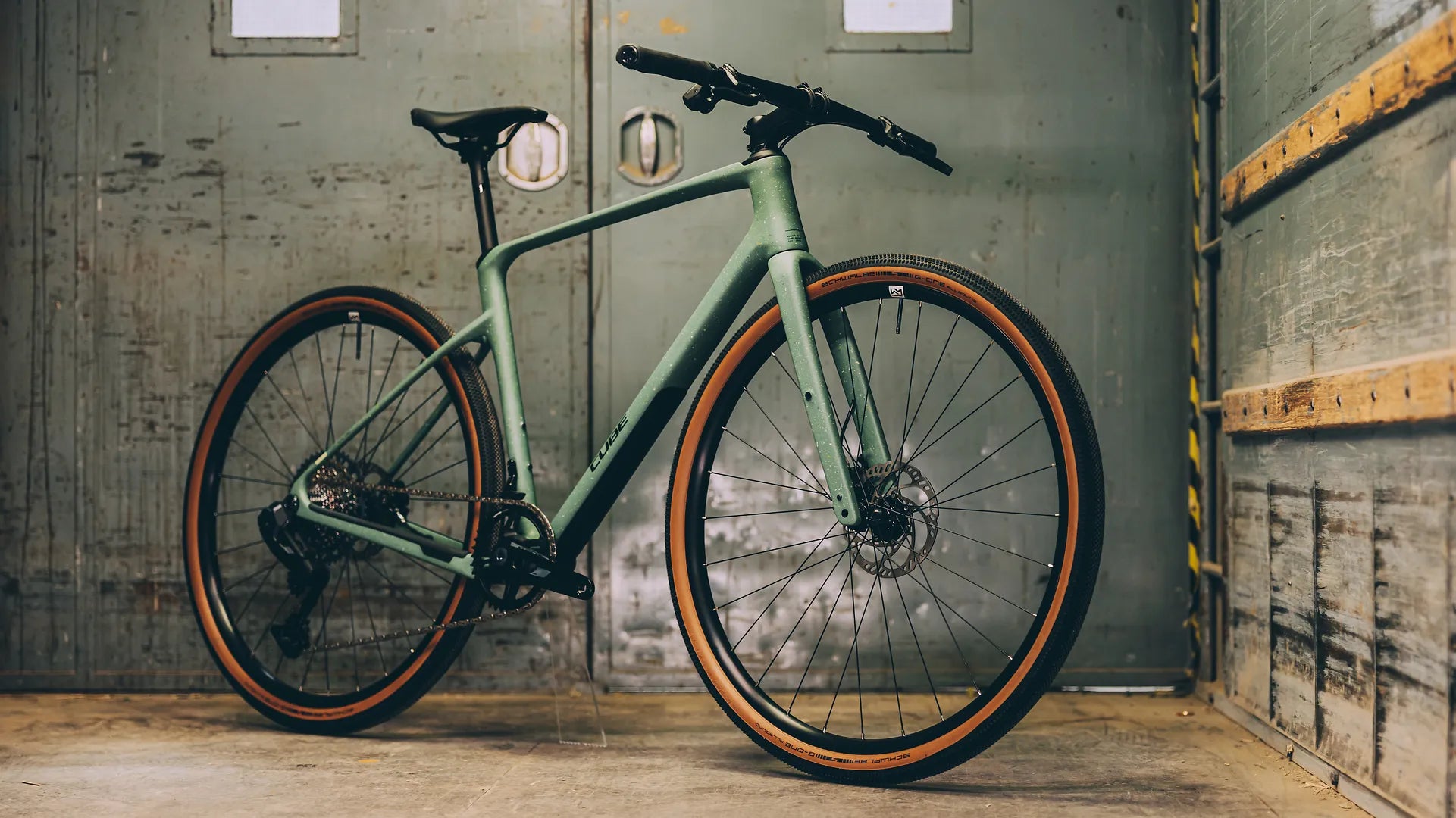 Grünes Cube NULANE C:62 SLT
, Gravelbike für Abenteuertouren, mit Scheibenbremsen und braunen Reifen, an grauer Wand gelehnt.