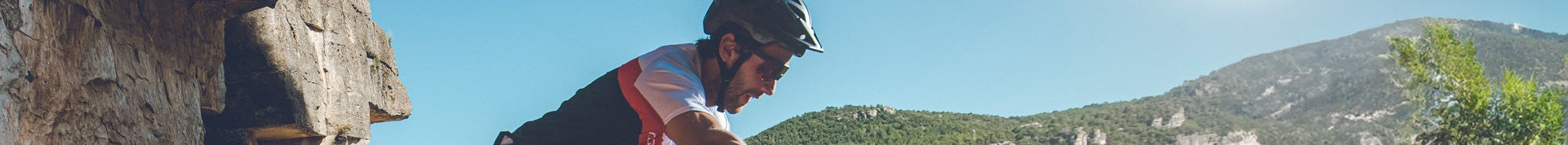 Ein Mountainbiker mit Helm fährt an einem steinigen Abhang entlang, umgeben von grünen Bergen und klarem Himmel.