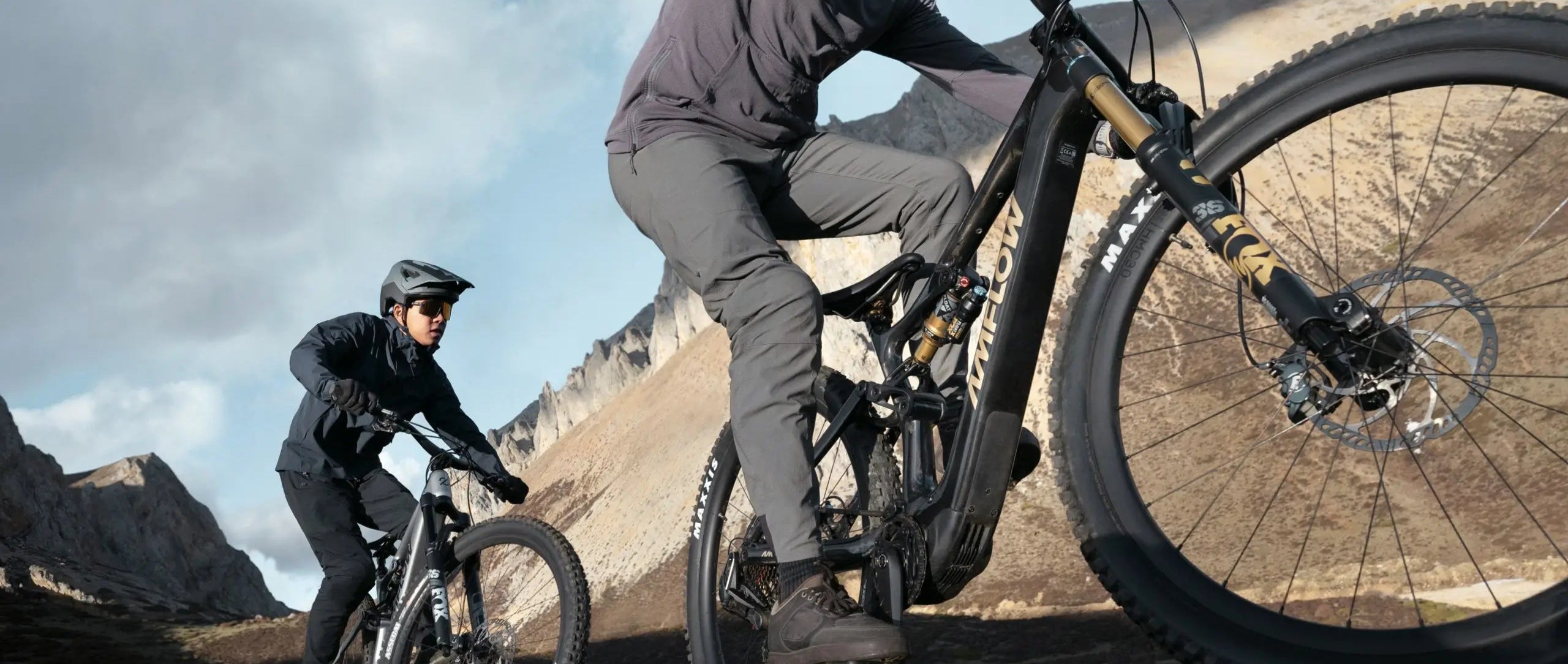 Zwei Personen fahren auf AMFLOW-Mountainbikes im Gebirge. Eines der Bikes hat FOX-Gabeln und MAXXIS-Reifen.